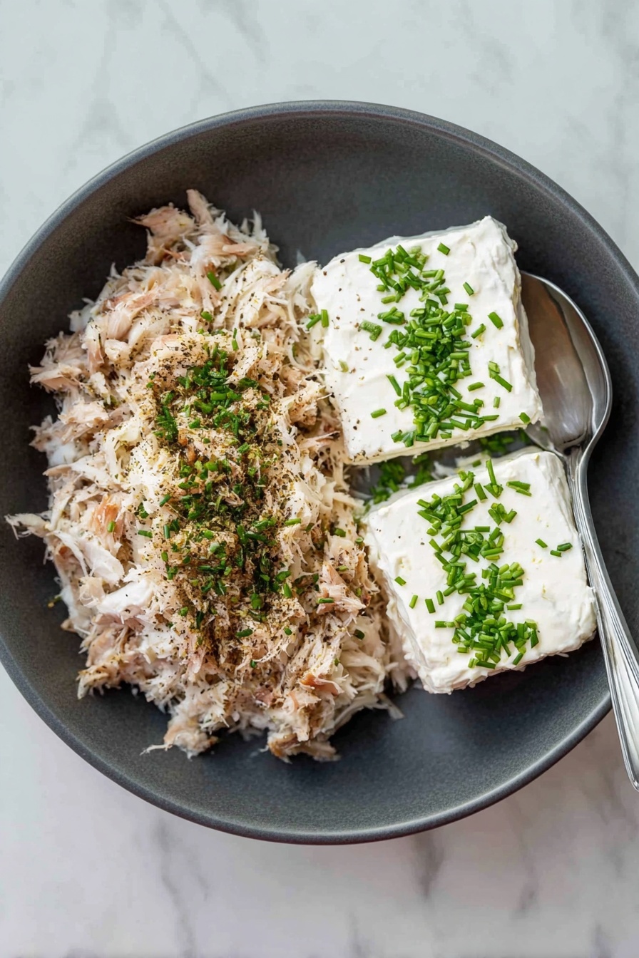 A dark grey bowl sits on a white marbled surface, filled with two main layers side by side: one side has a large mound of light brown shredded fish with a sprinkle of black pepper and chopped green herbs on top, while the other side shows two rectangular blocks of smooth white cream cheese topped with finely chopped green herbs. A silver spoon rests inside the bowl on the right edge. photo taken with an iphone --ar 2:3 --v 7